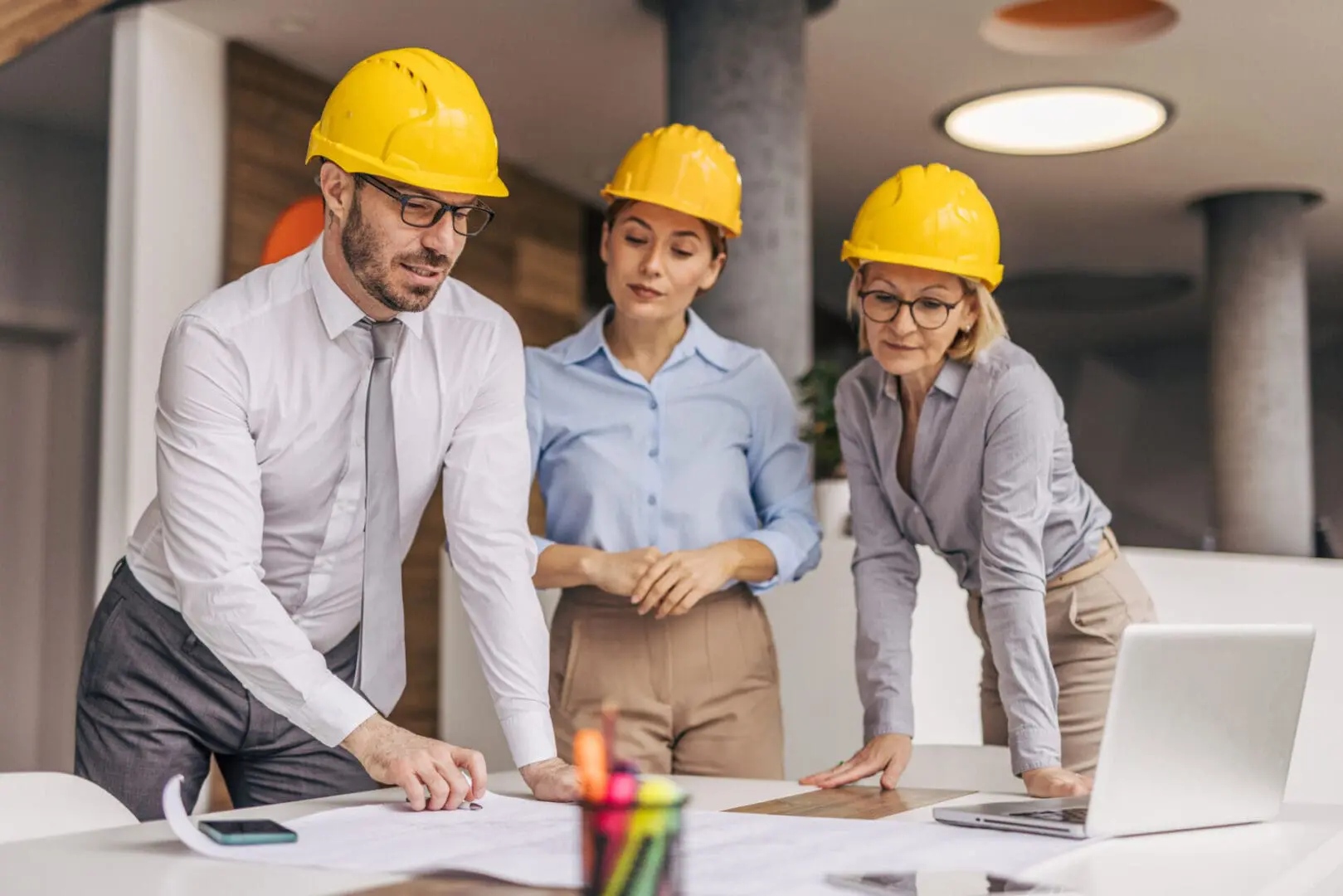 Three architects, wearing hardhats, collaborating on a project while examining blueprints and utilizing a laptop in a modern office environment, showcasing teamwork and expertise
